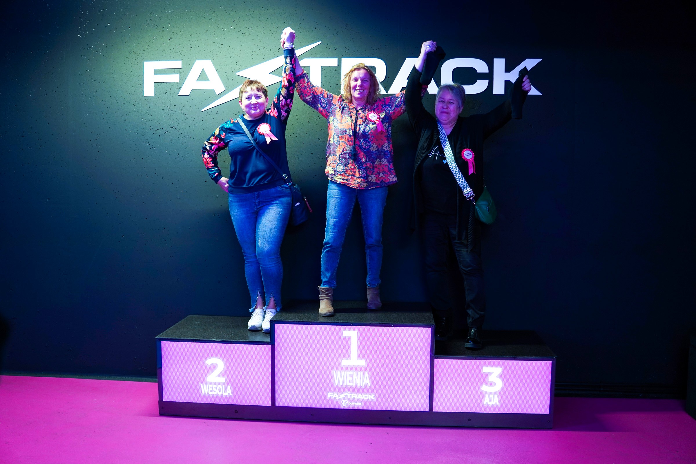 Trzy kobiety stoją na podium toru Fast Track, trzymając się za ręce w geście zwycięstwa. Za nimi ciemna ściana z logo toru.